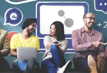 Group of people sitting on the floor in front of a decorated wall. They each hold electronic devices such as phones, laptops, or tablets.